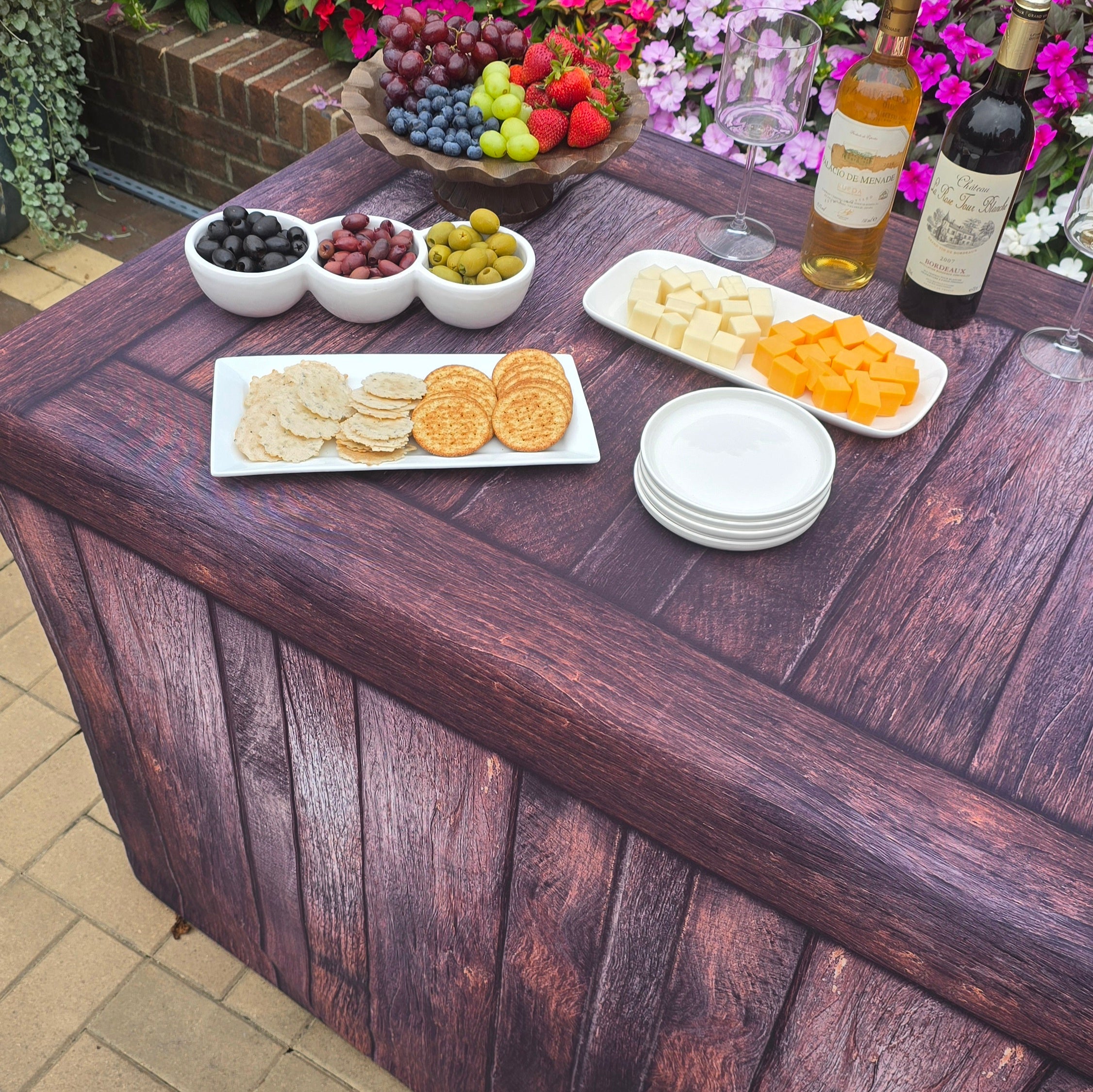 A fabric fitted table cover that has a wood look design on a folding table outside by a pool with catered food and charcuterie, wine and glasses.